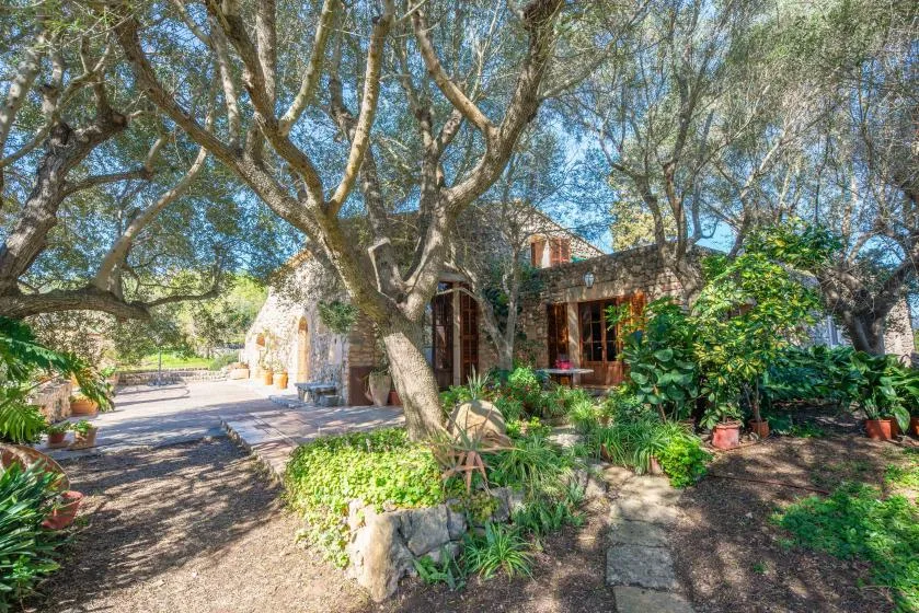 A stone house stands among mature trees and leafy garden plants.