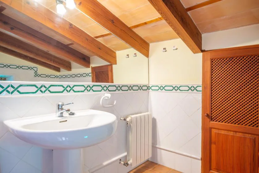 A tiled bathroom with a wall-mounted sink, radiator, and exposed wooden ceiling beams.