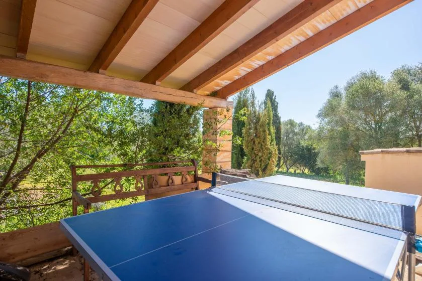 A blue table tennis table sits on a covered outdoor patio surrounded by trees.