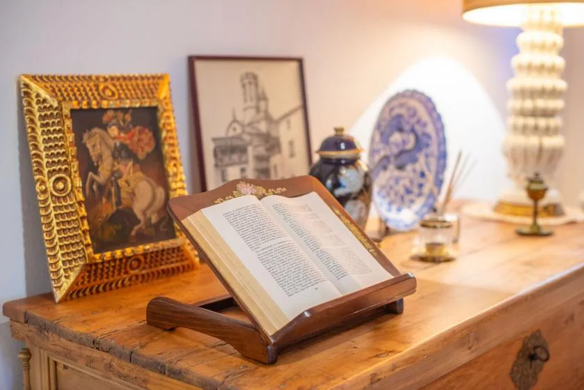 An open book rests on a wooden stand atop a table beside framed artwork.