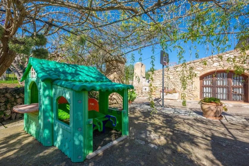 A colorful outdoor playhouse sits in a sunny courtyard beneath leafy trees.