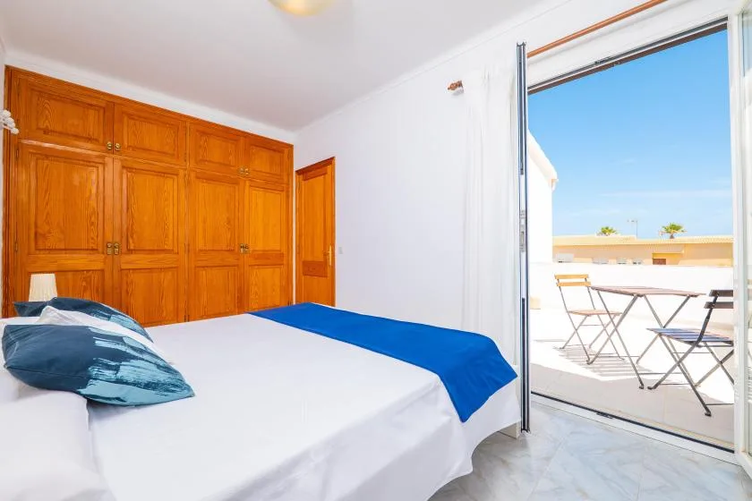 A bedroom with a bed beside an open sliding glass door to a sunny terrace.