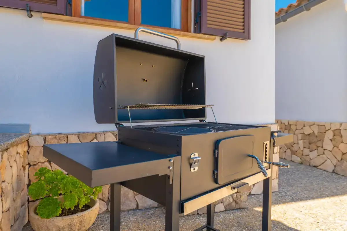 An outdoor barbecue grill stands beside a white wall with shuttered windows.