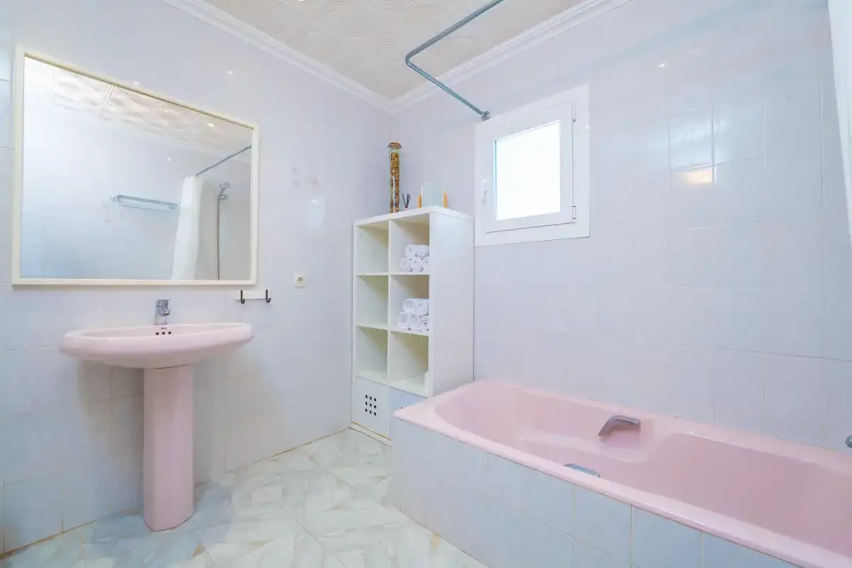 A pale tiled bathroom with a pink bathtub and pedestal sink.
