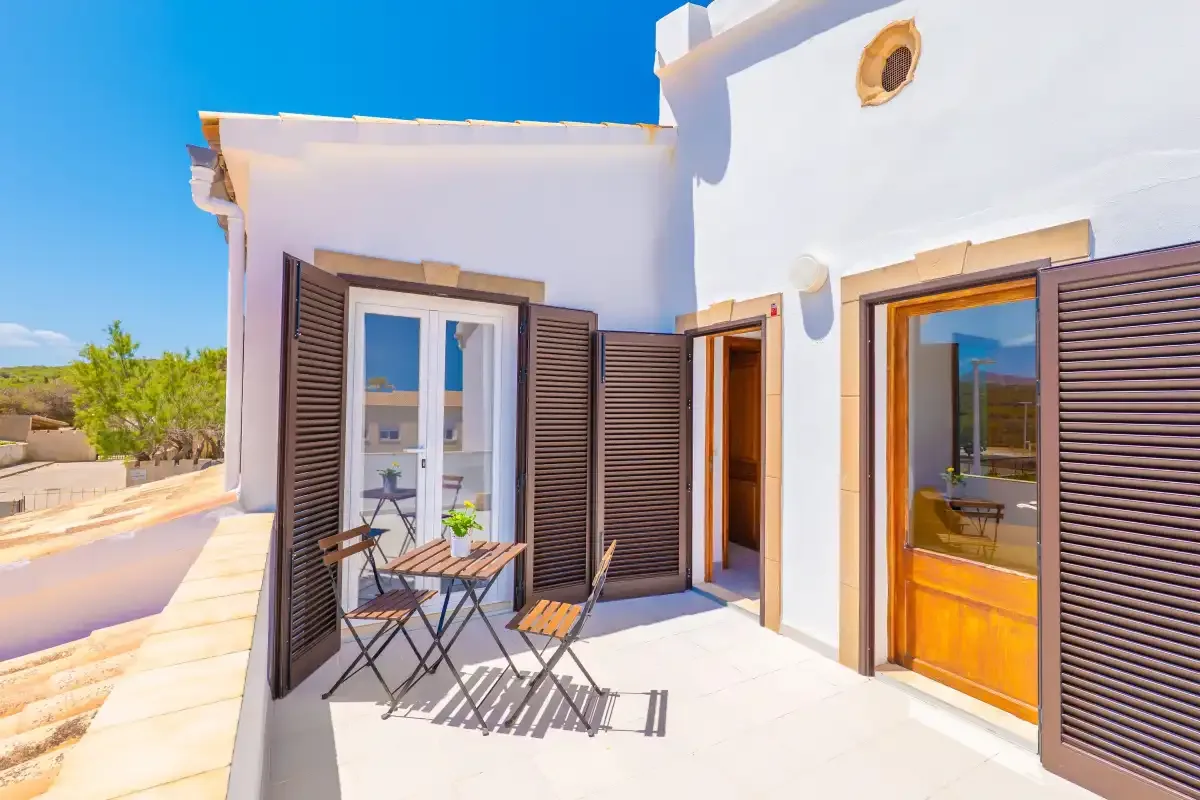 A sunlit white terrace with dark shutters, glass doors, and a small table with chairs.