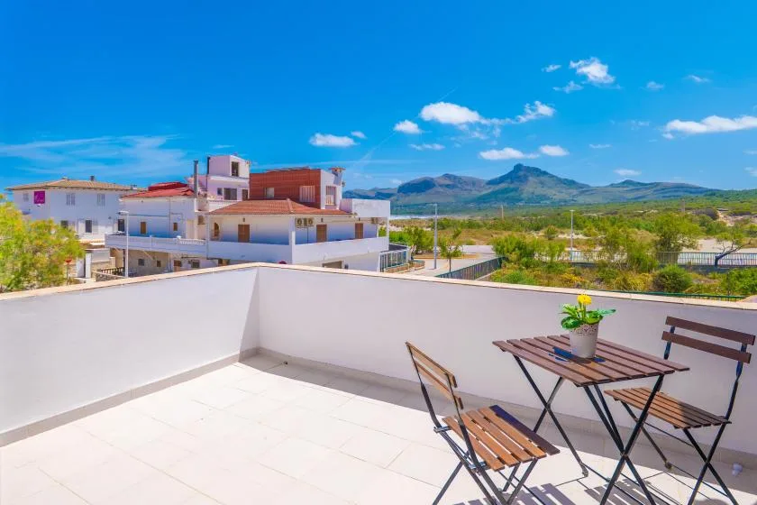 A sunlit rooftop terrace has a small table and seating overlooking nearby buildings and mountains.