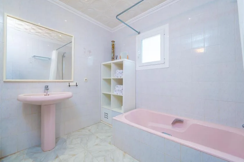 A tiled bathroom with a pink bathtub, pedestal sink, and wall shelves.