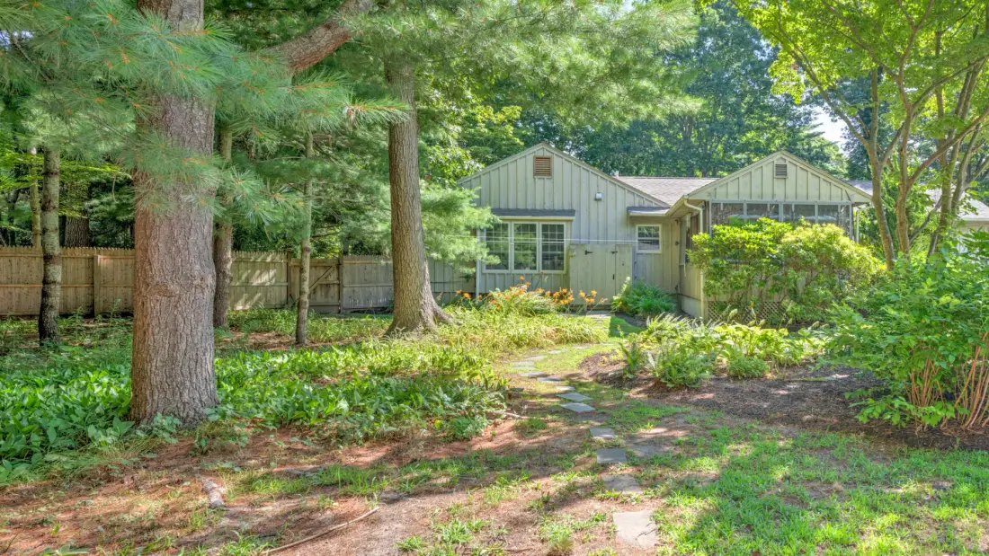 A small house stands among trees and greenery beside a garden path.