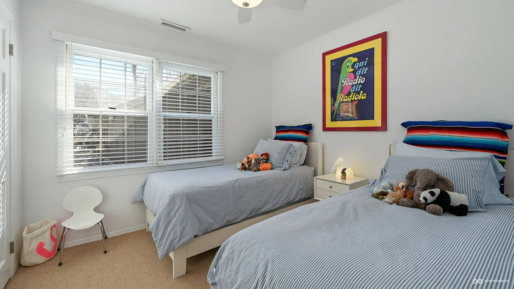 A bright bedroom with two beds, striped pillows, and windows with blinds.