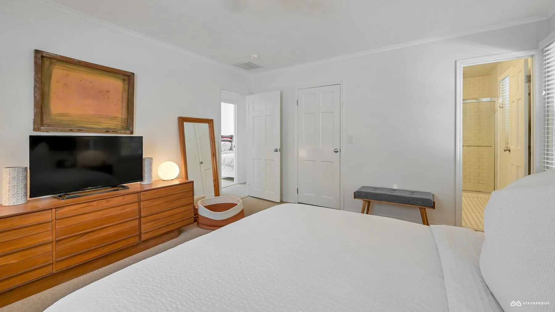 A bright bedroom with a bed, dresser, television, and an open doorway to a bathroom.