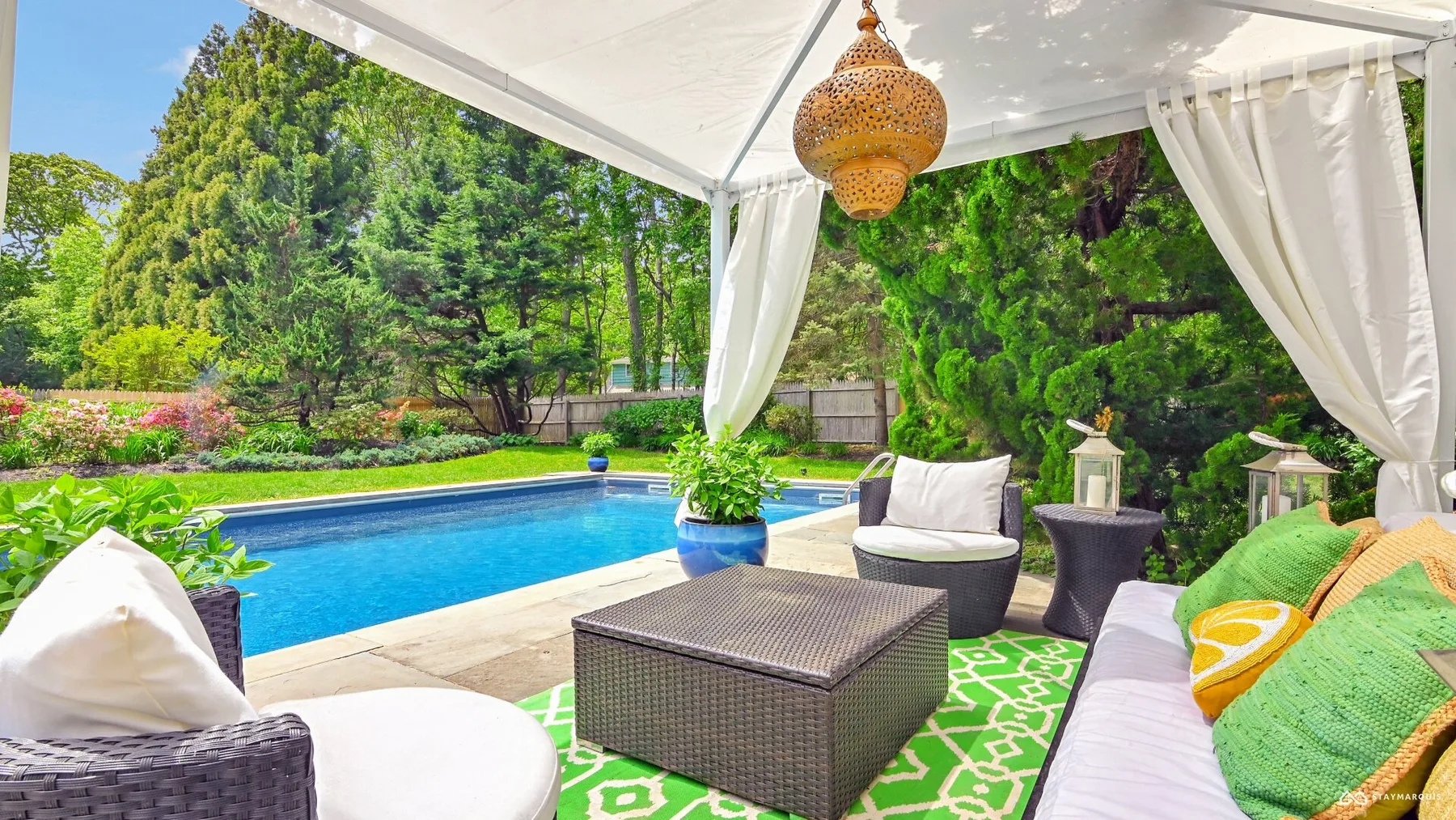 A covered outdoor seating area beside a swimming pool, with cushions and greenery.