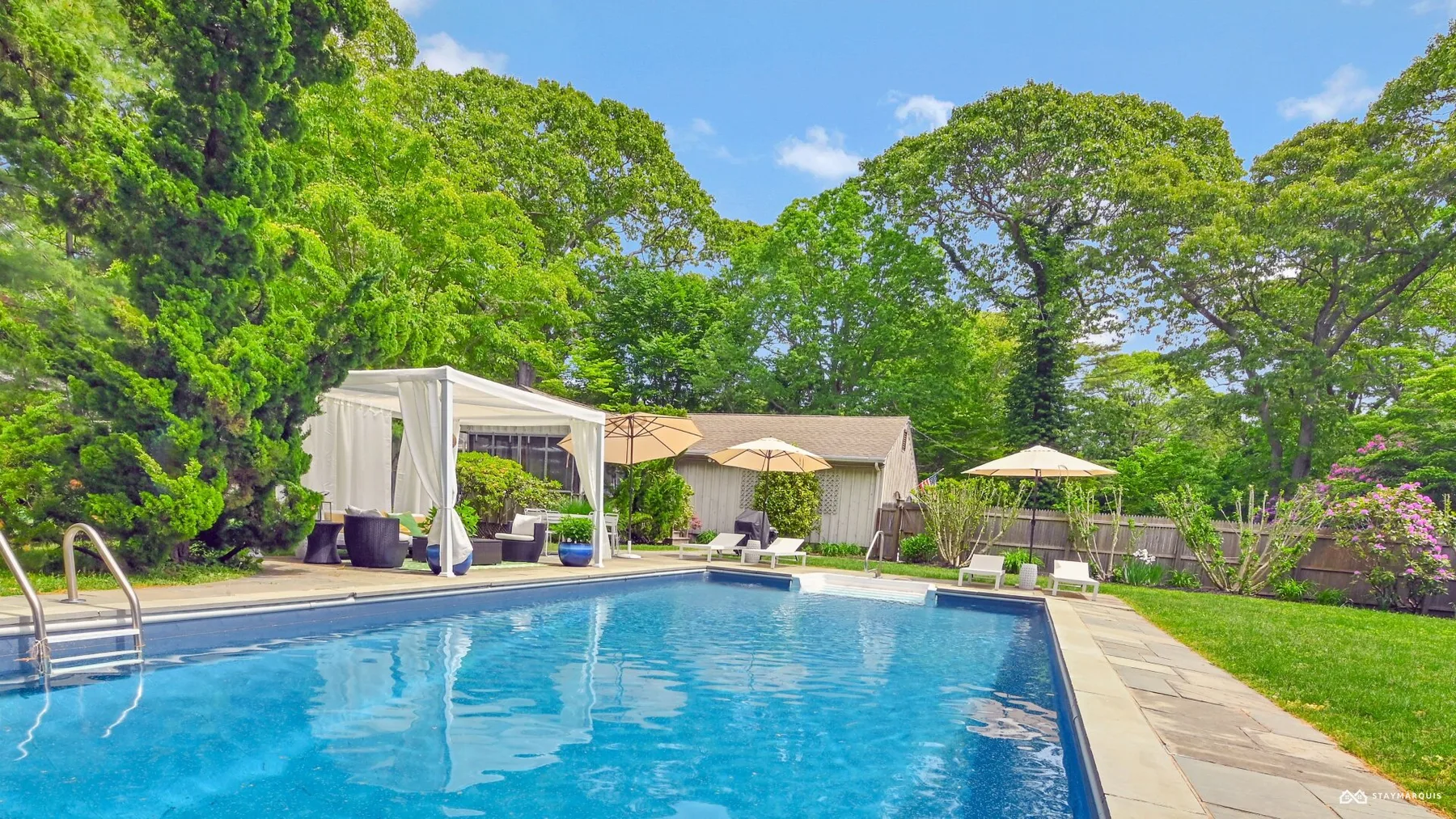 A swimming pool sits beside a paved patio and leafy trees.
