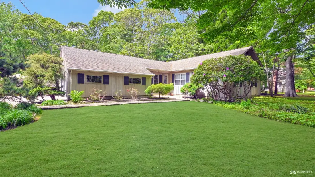 A single-story house stands behind a broad green lawn, surrounded by trees.