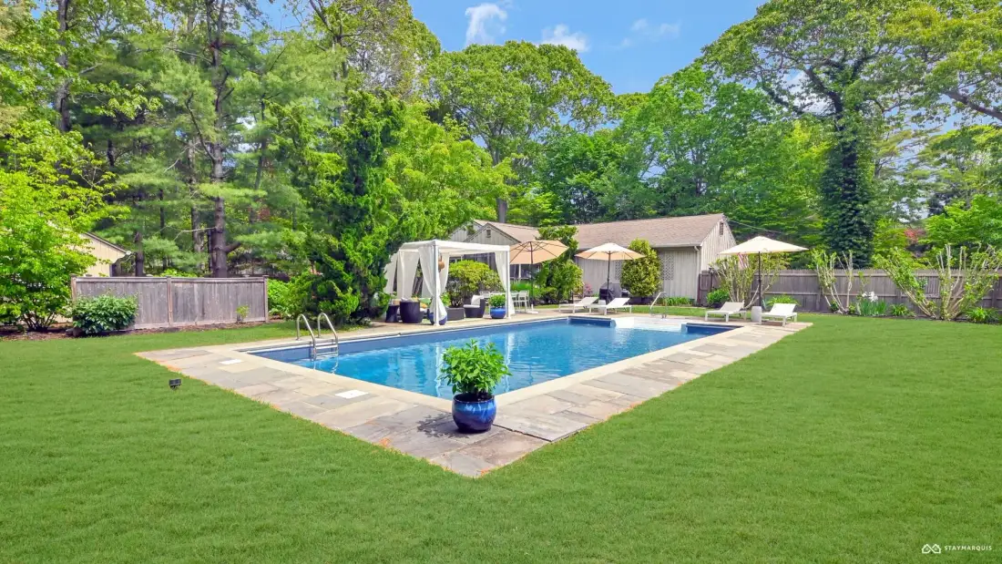 A backyard pool is surrounded by a lawn, trees, and outdoor seating.