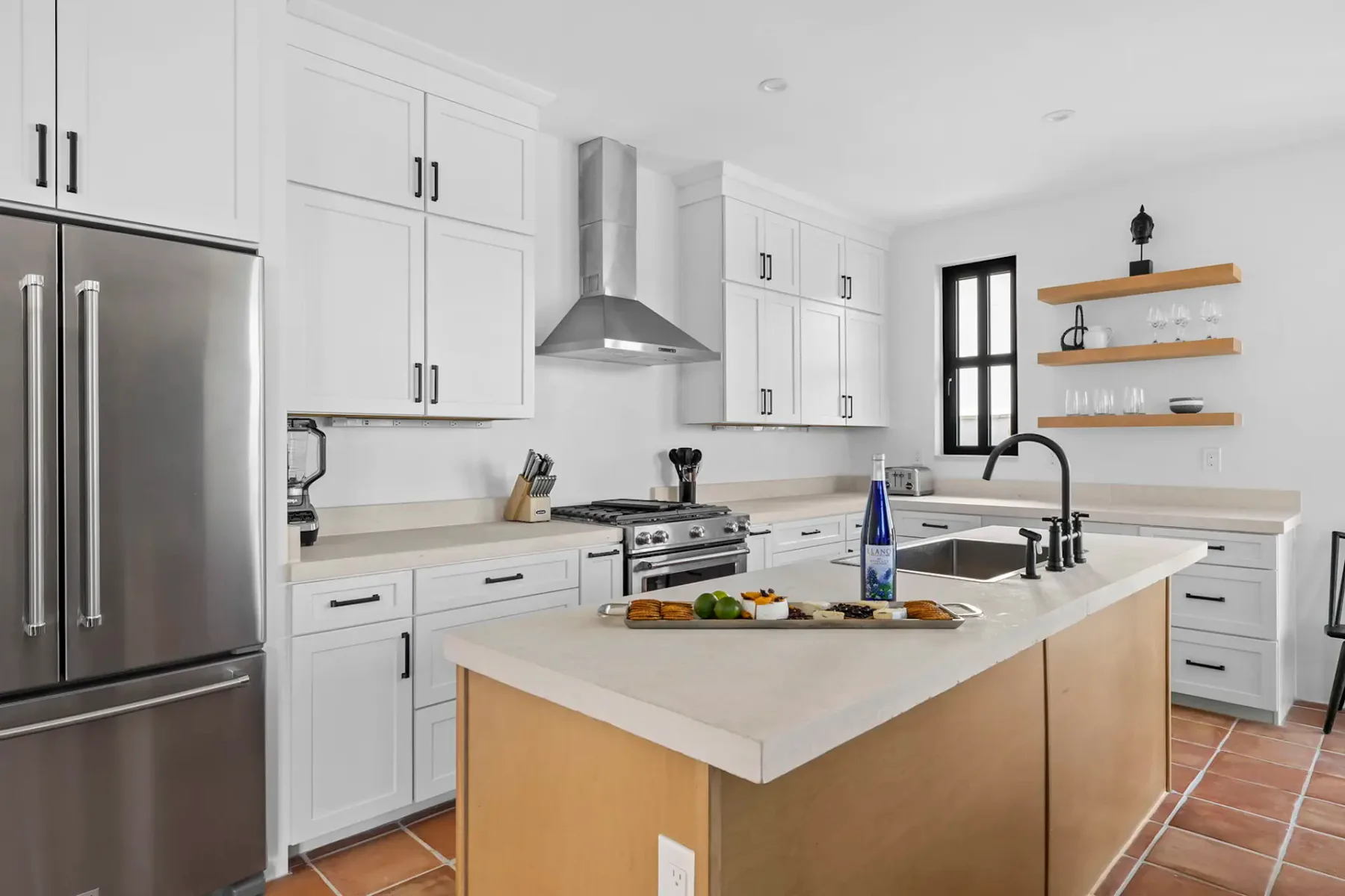 Cocina moderna con gabinetes blancos, electrodomésticos de acero inoxidable y una isla central con fregadero y una tabla de cortar con alimentos.