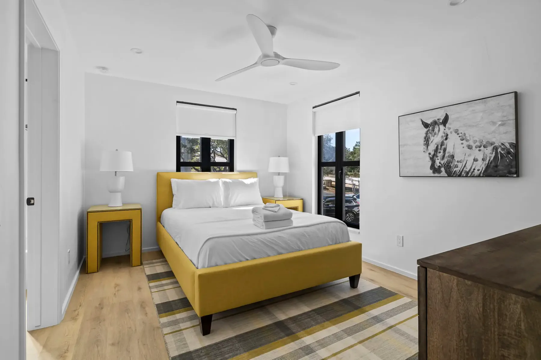 Dormitorio minimalista con una cama amarilla, una alfombra a rayas y una obra de arte de un caballo. Dos ventanas, mesas de noche con lámparas y un ventilador de techo completan la decoración.