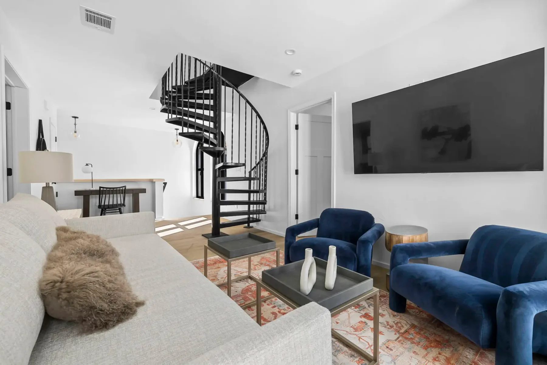Modern living room with a gray sofa, blue armchairs, spiral staircase, large TV, and a desk in the background. Neutral tones and minimal decor.