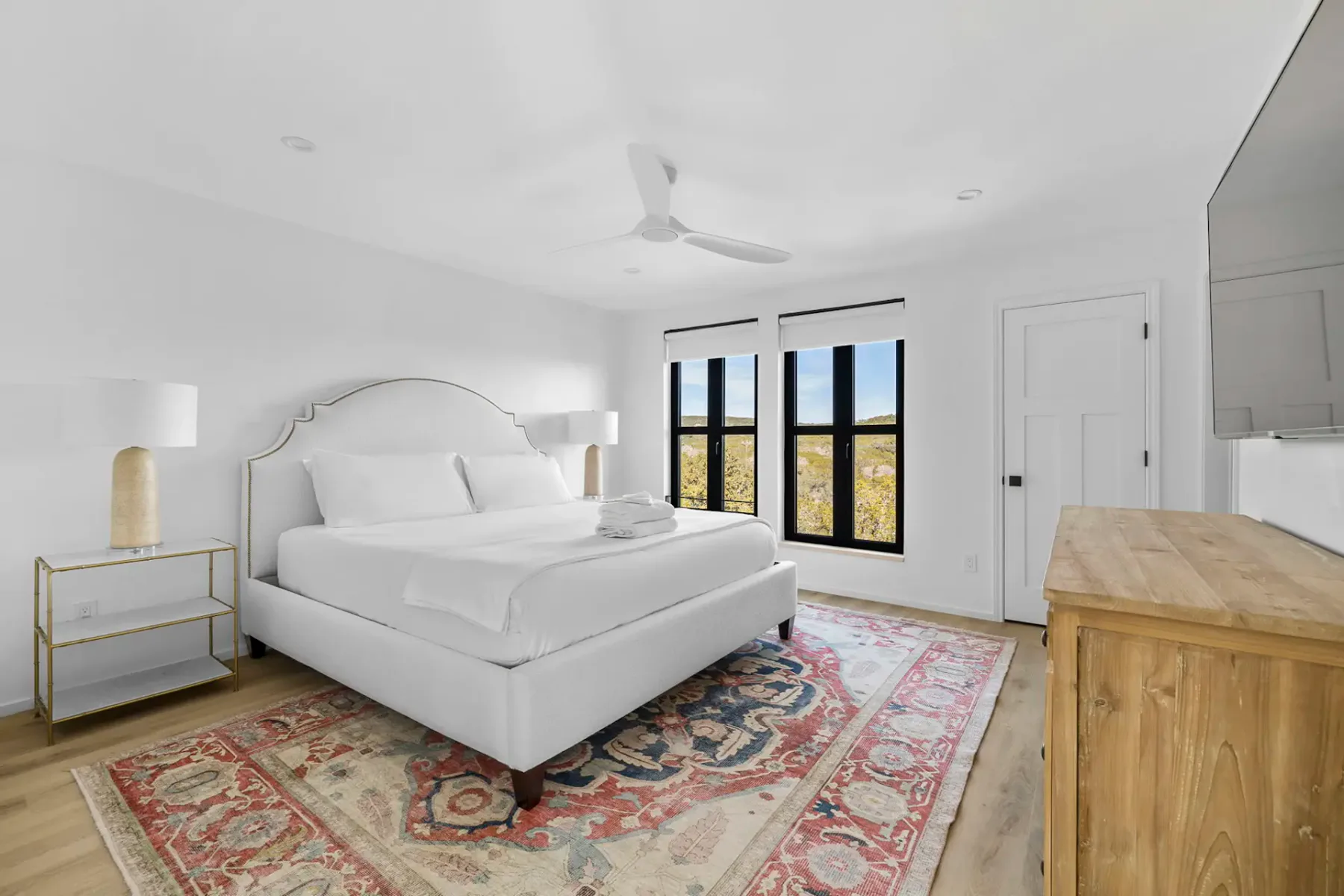 Un dormitorio luminoso con una cama blanca, una alfombra estampada y ventanas con marcos negros.