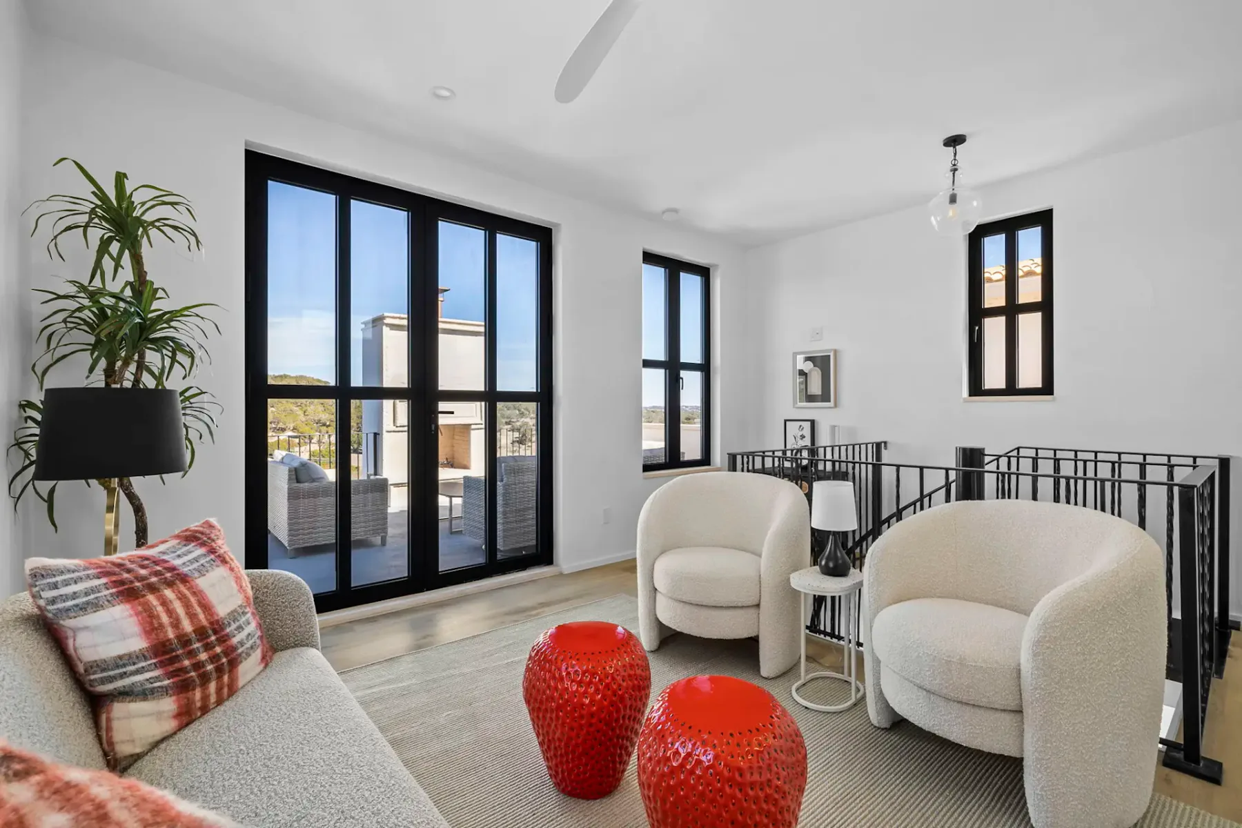 Sala de estar moderna con paredes blancas, ventanas grandes, dos sillones blancos redondos, un sofá con cojines a cuadros y objetos decorativos rojos sobre una alfombra.