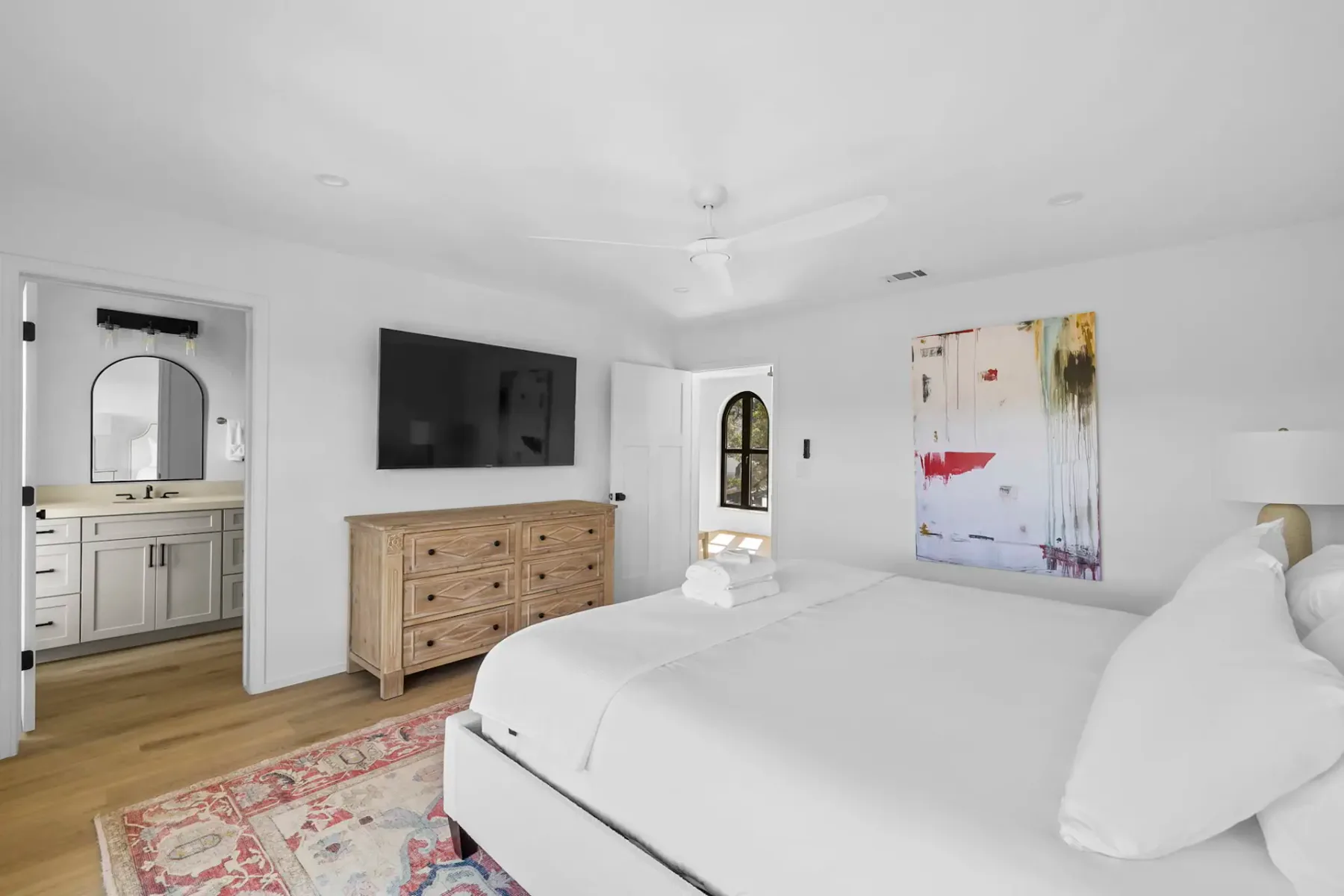 Dormitorio luminoso con una cama grande, una cómoda de madera, arte en la pared, un ventilador de techo y un televisor montado en la pared. Una puerta da acceso a un baño contiguo.