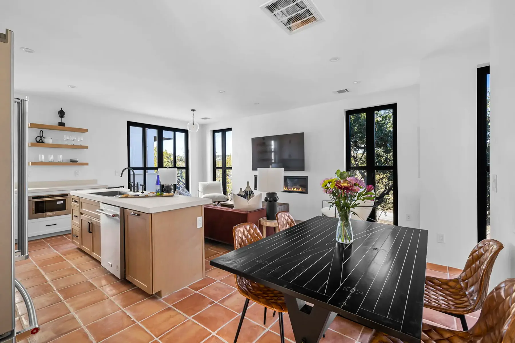 Modern open-plan kitchen and living area with terracotta tile floor, black dining table, wicker chairs, and vase of flowers near large windows.