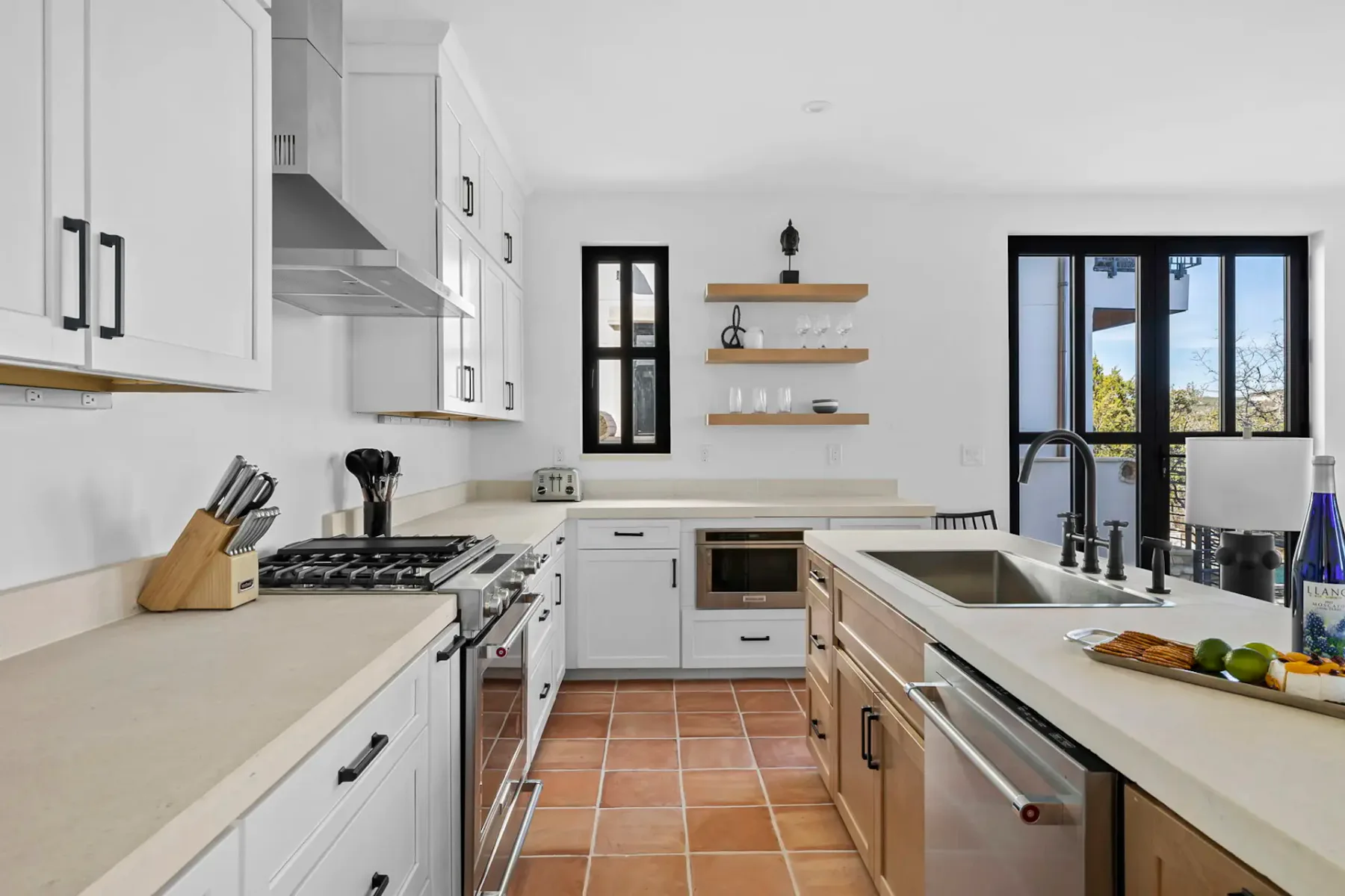 Modern kitchen with white cabinets, stainless steel appliances, black fixtures, and terracotta floor tiles. Open shelves display cups and decor.