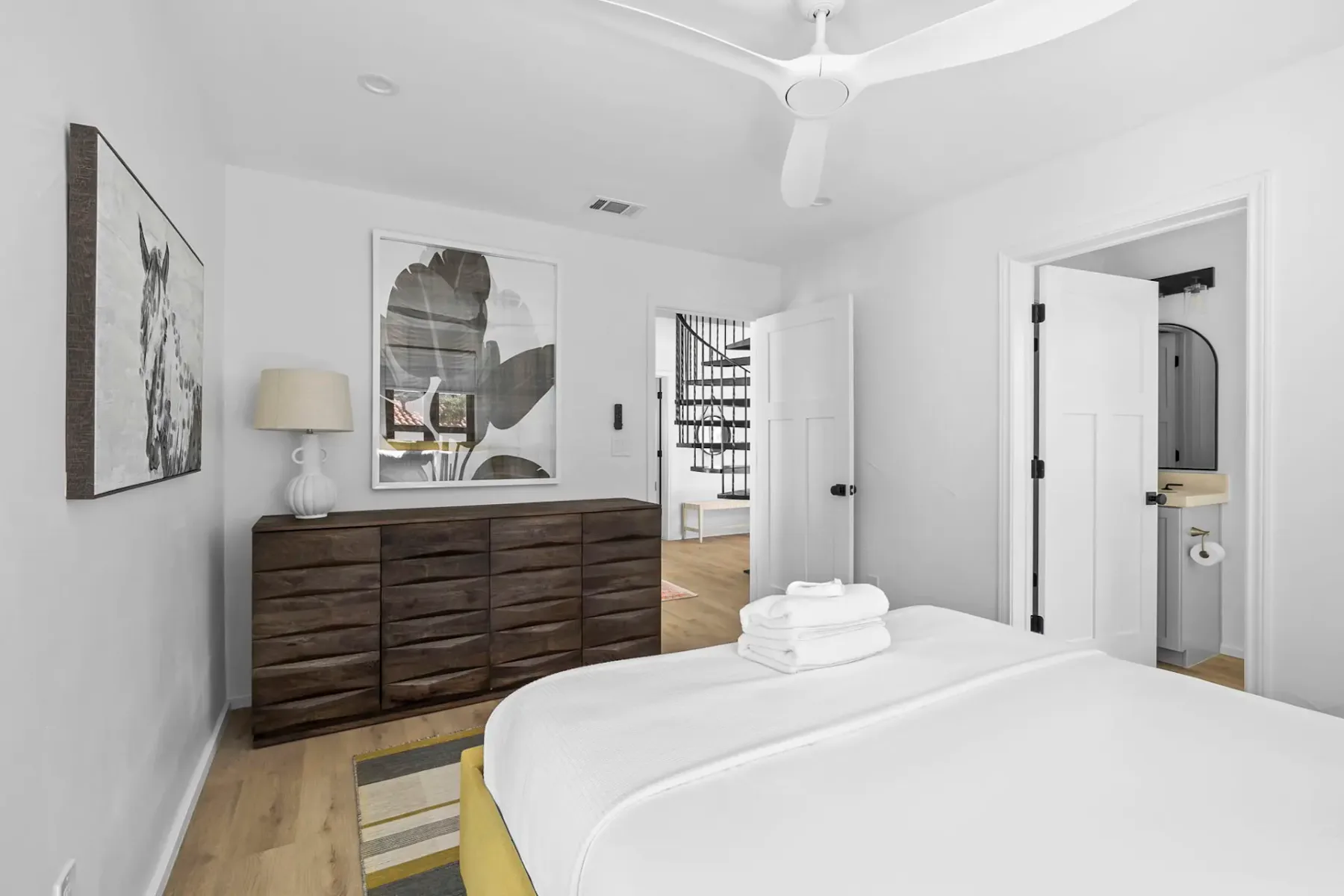 Dormitorio minimalista con una cama grande, una cómoda de madera, láminas artísticas, un ventilador de techo y una vista de la puerta abierta de un baño. Predominan el blanco y los tonos neutros.