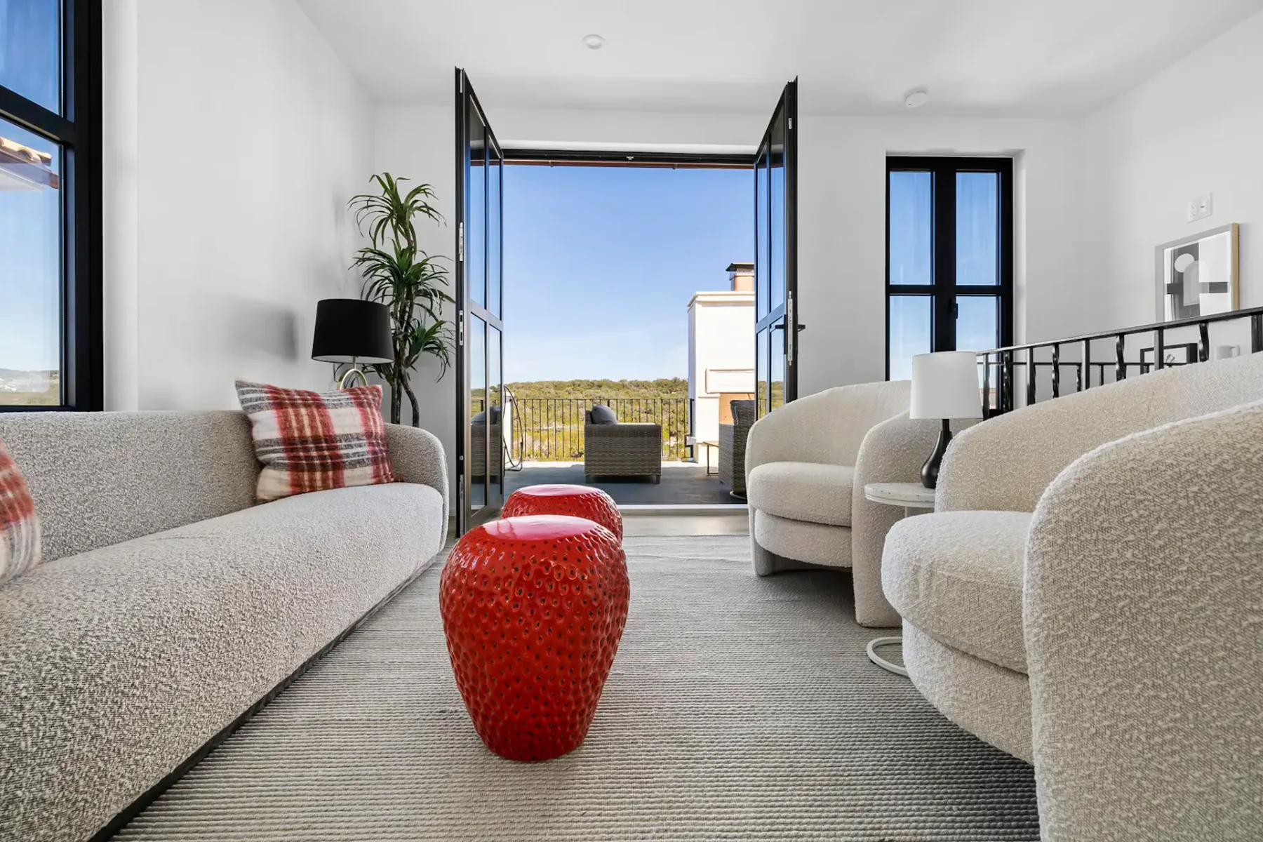 Modern living room with a gray sofa, round armchairs, red textured stools, and open doors leading to a balcony with a scenic view.