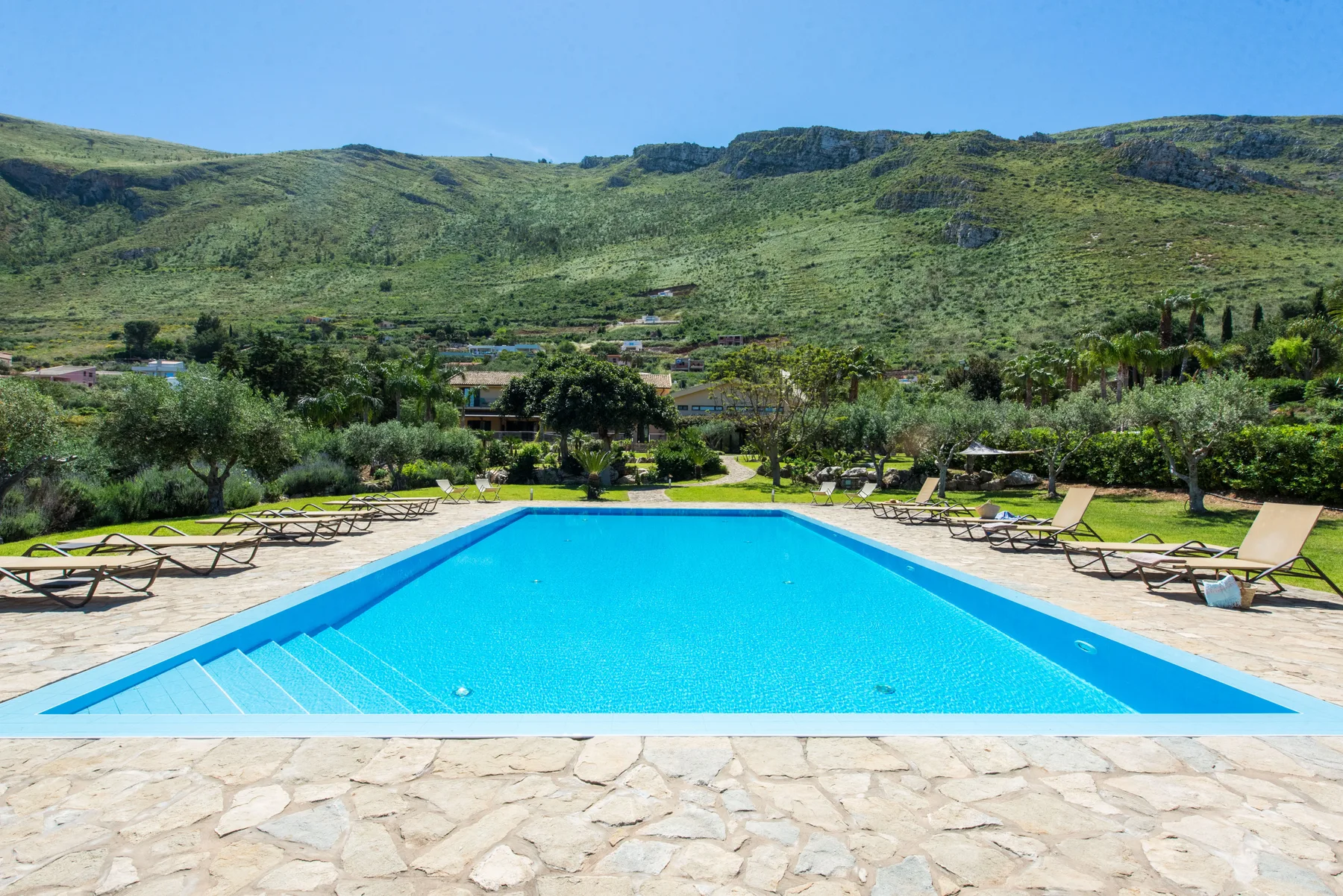 An outdoor swimming pool is surrounded by sun loungers and green hills.
