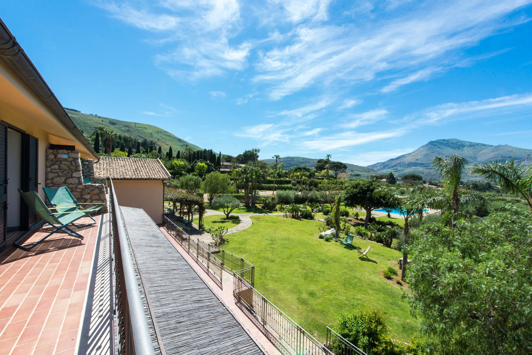 A garden and swimming pool sit below a mountain view beside a building terrace.