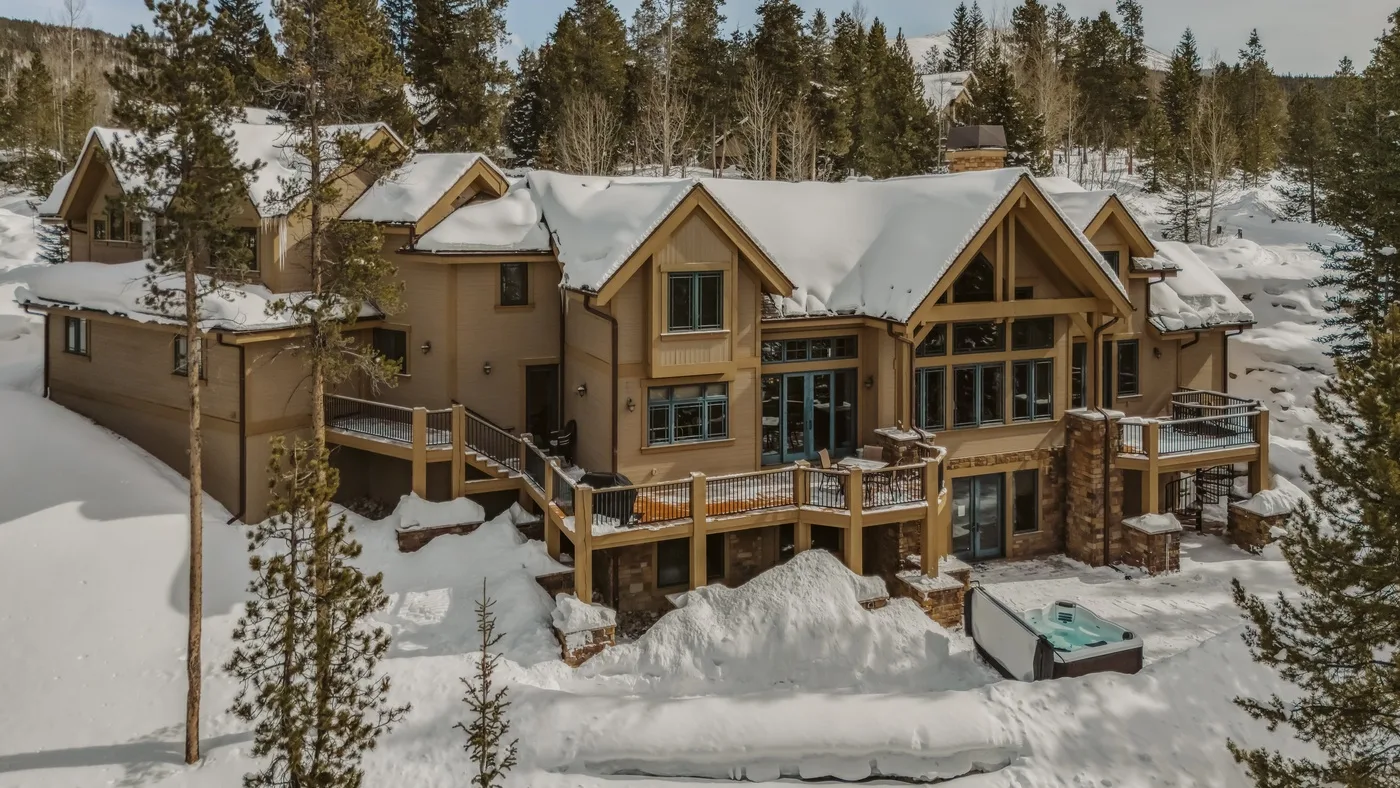 Un gran alojamiento está en un bosque nevado, con una terraza y una bañera de hidromasaje exterior.