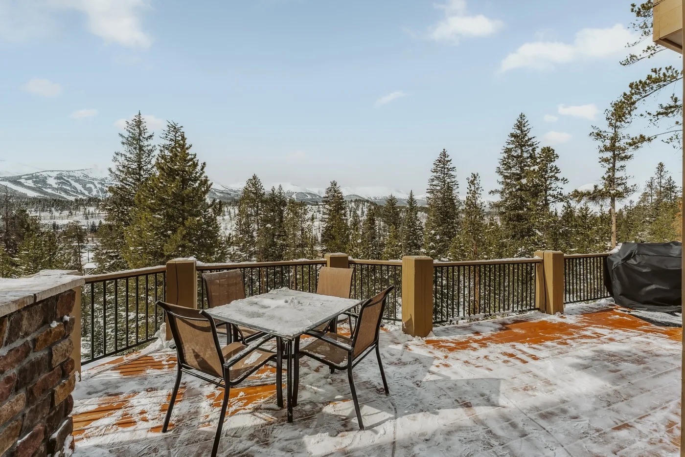 Una terraza exterior con nieve, mesa y asientos ofrece vistas a un paisaje boscoso.