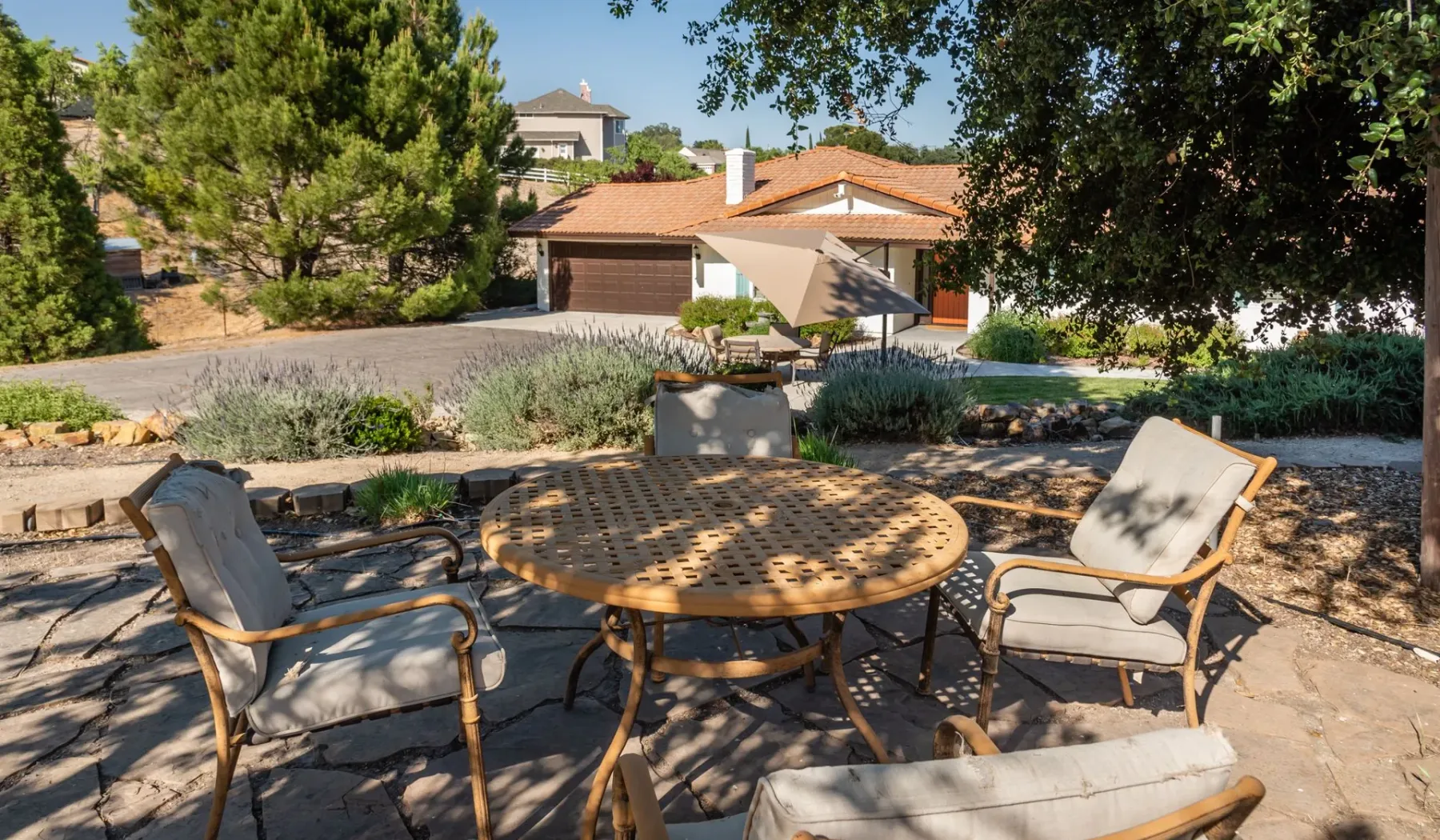 Un patio al aire libre con una mesa redonda y asientos acolchados bajo árboles frondosos.