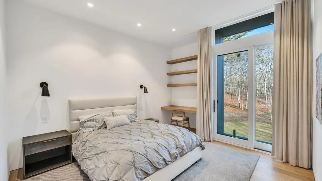 Un dormitorio luminoso con una cama junto a grandes puertas de vidrio y vistas a un bosque.