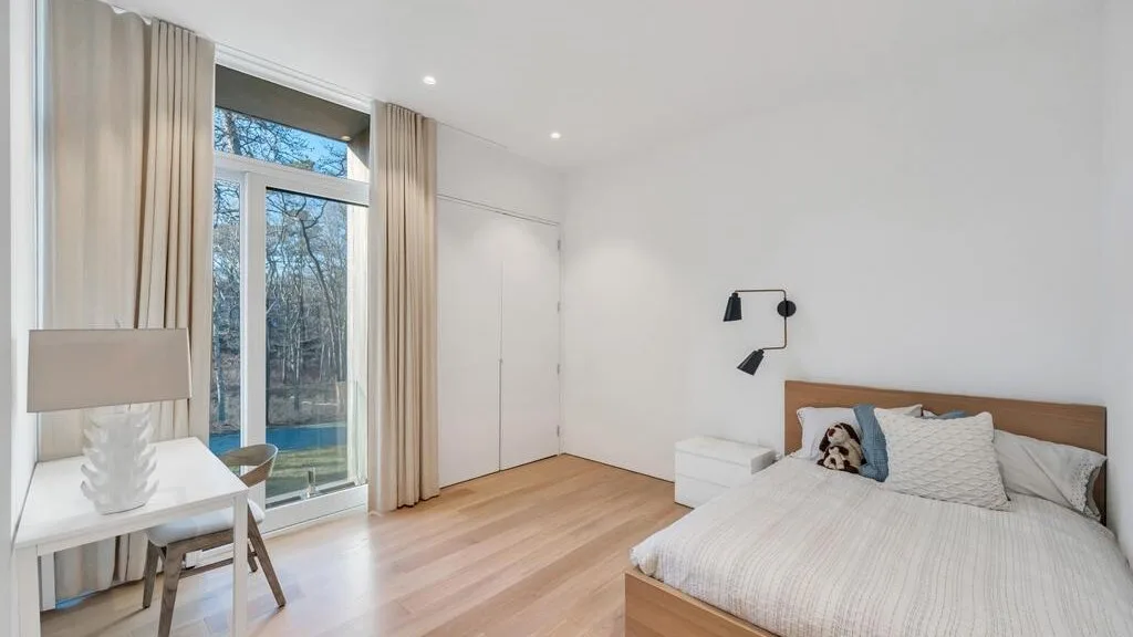 Un dormitorio luminoso con una cama junto a ventanas altas y cortinas claras.