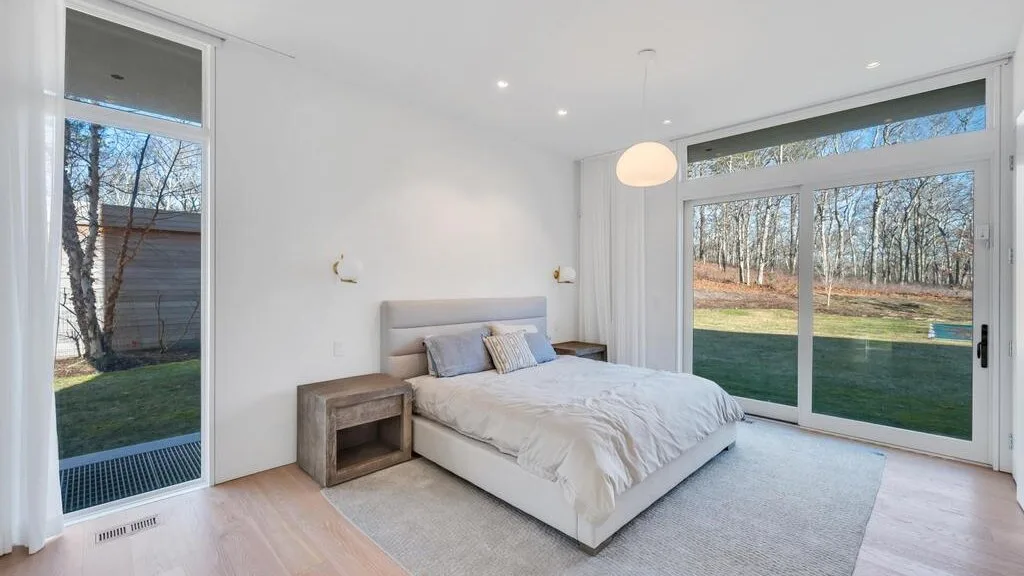 Un dormitorio luminoso con una cama junto a grandes ventanas que dan a un césped y árboles.