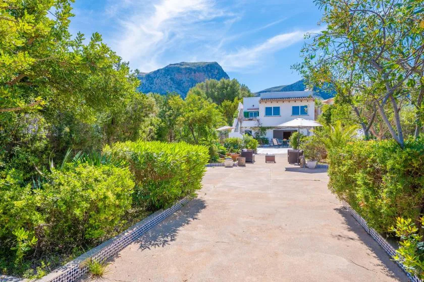 A paved garden path leads toward a white building, with trees and mountains behind it.
