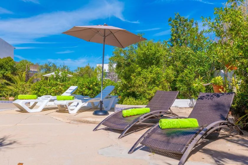 An outdoor poolside patio with reclining loungers, an umbrella, and surrounding greenery.