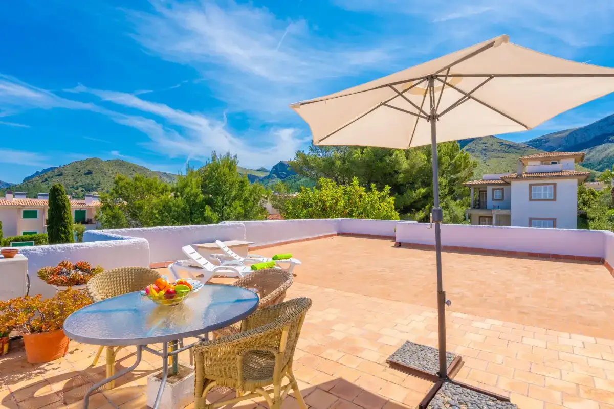 A sunny rooftop terrace has a dining table, lounge seating, and a large umbrella.