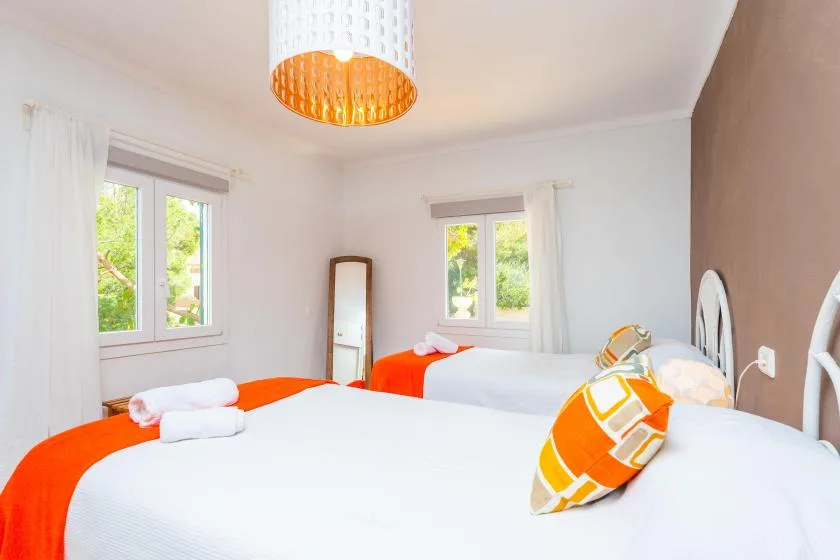 A bright bedroom with white bedding, orange accents, and windows overlooking greenery.