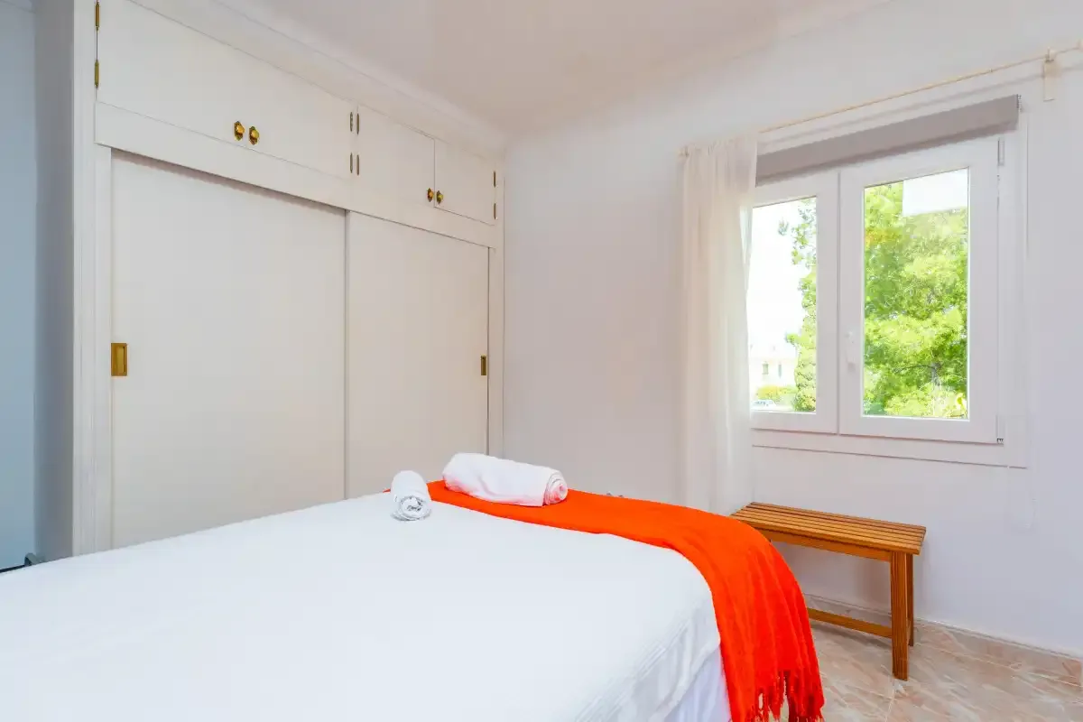 A bedroom with white bedding, a bright orange throw, and a window overlooking greenery.