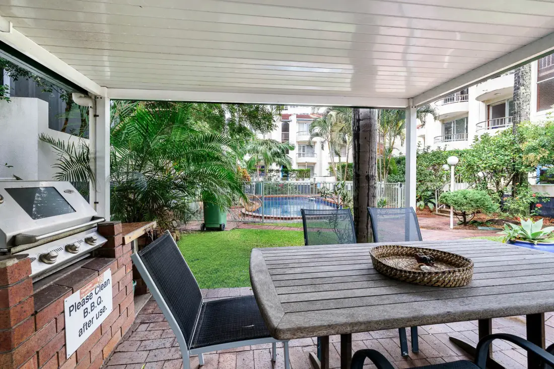 A covered patio with a table and seating overlooks a garden and swimming pool.