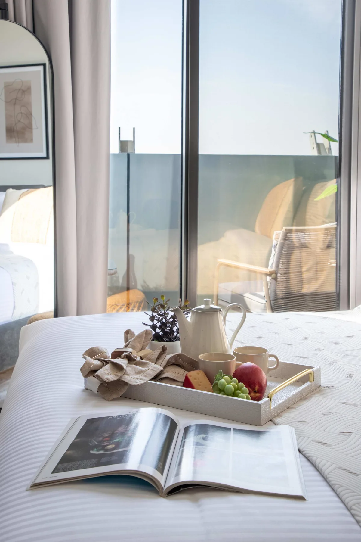 Un dormitorio luminoso con una cama, un libro abierto y una bandeja junto a la ventana.