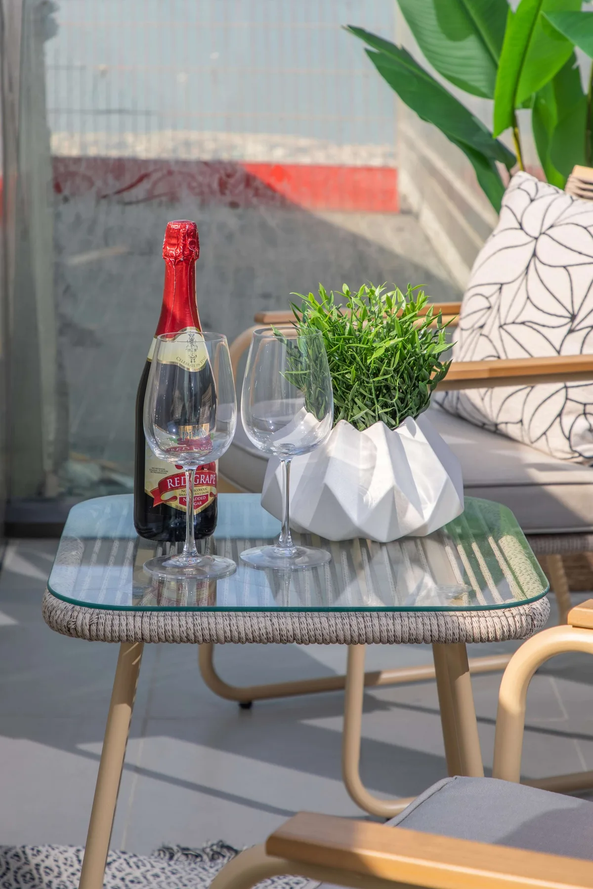 Una zona de estar en el patio con una mesa de vidrio, una botella de vino, copas y una planta.