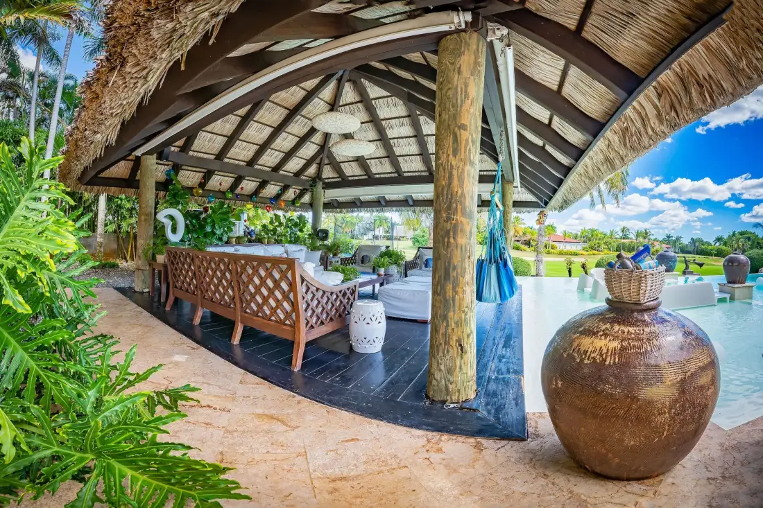 Un salón abierto junto a la piscina está bajo un techo de paja, junto a plantas tropicales.