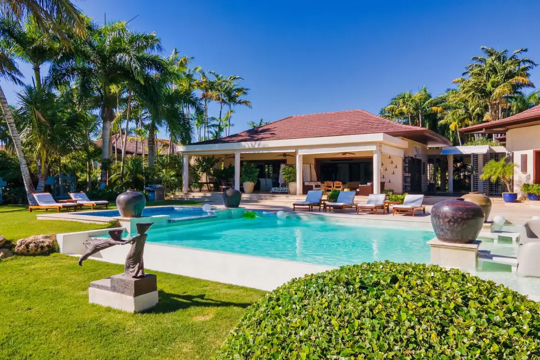 Una piscina tropical al aire libre está junto a un pabellón con techo de tejas y vegetación.