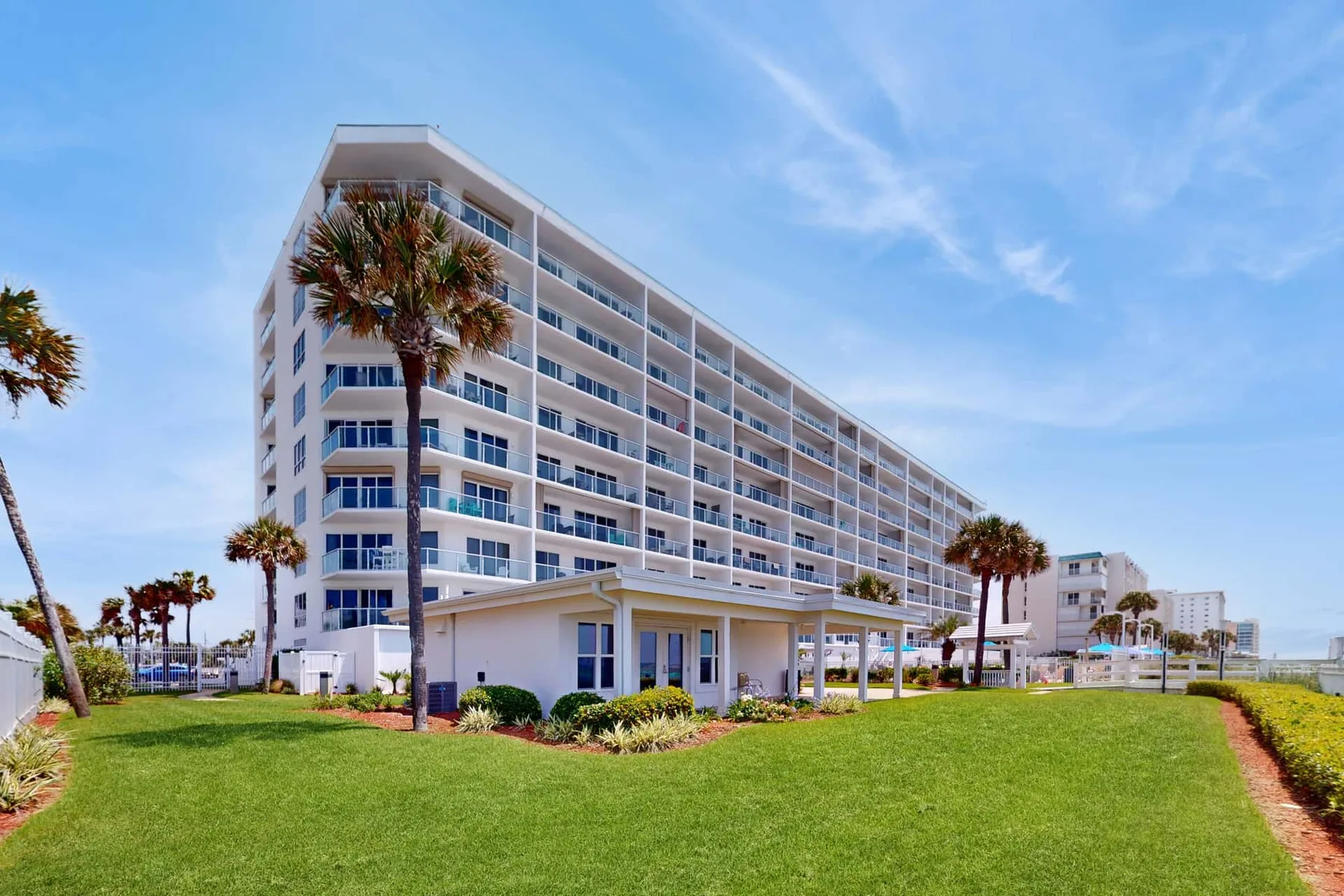 A large white multi-story building stands behind a grassy lawn and palm trees.