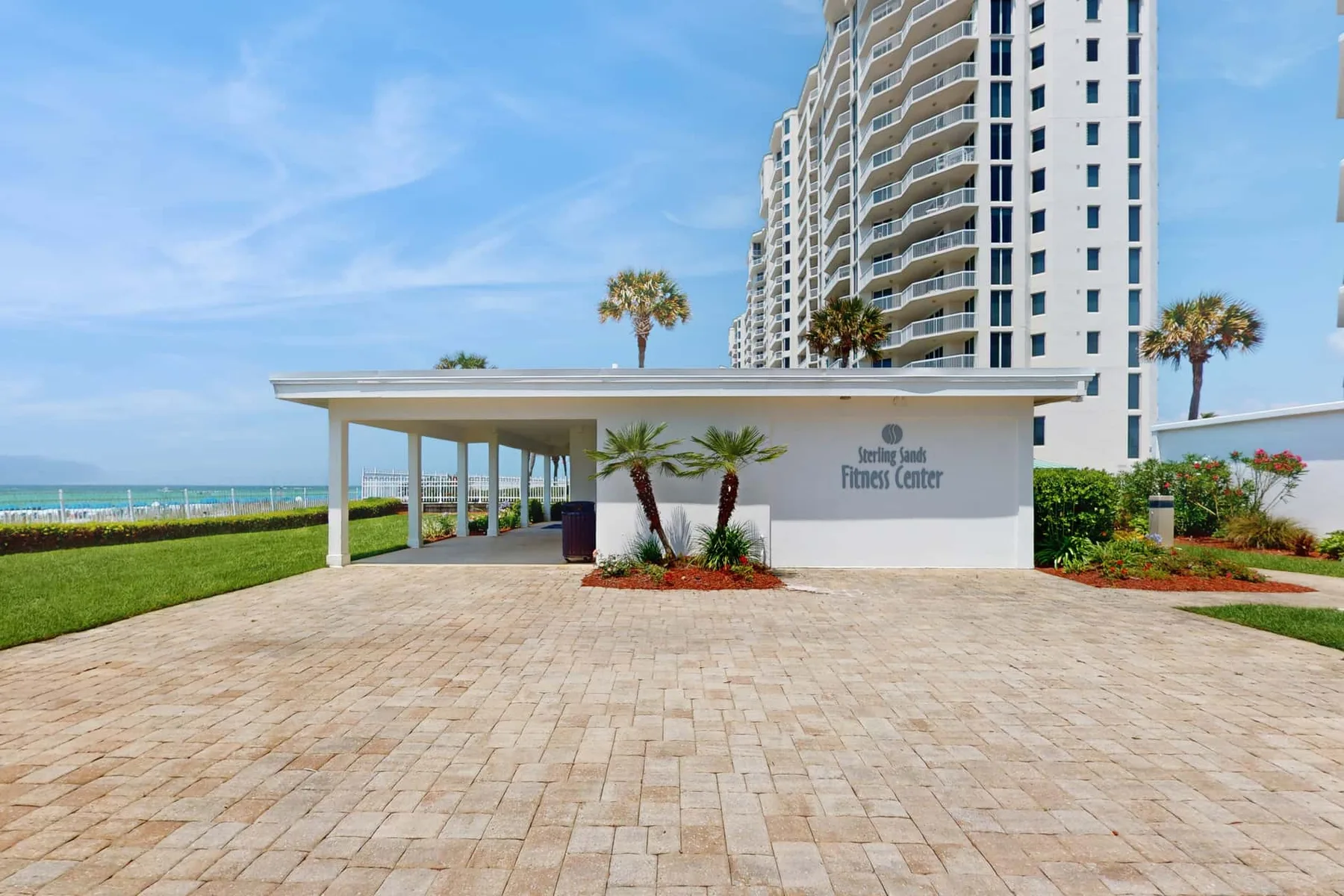 A paved entrance courtyard fronts a covered walkway beside a beachfront high-rise.