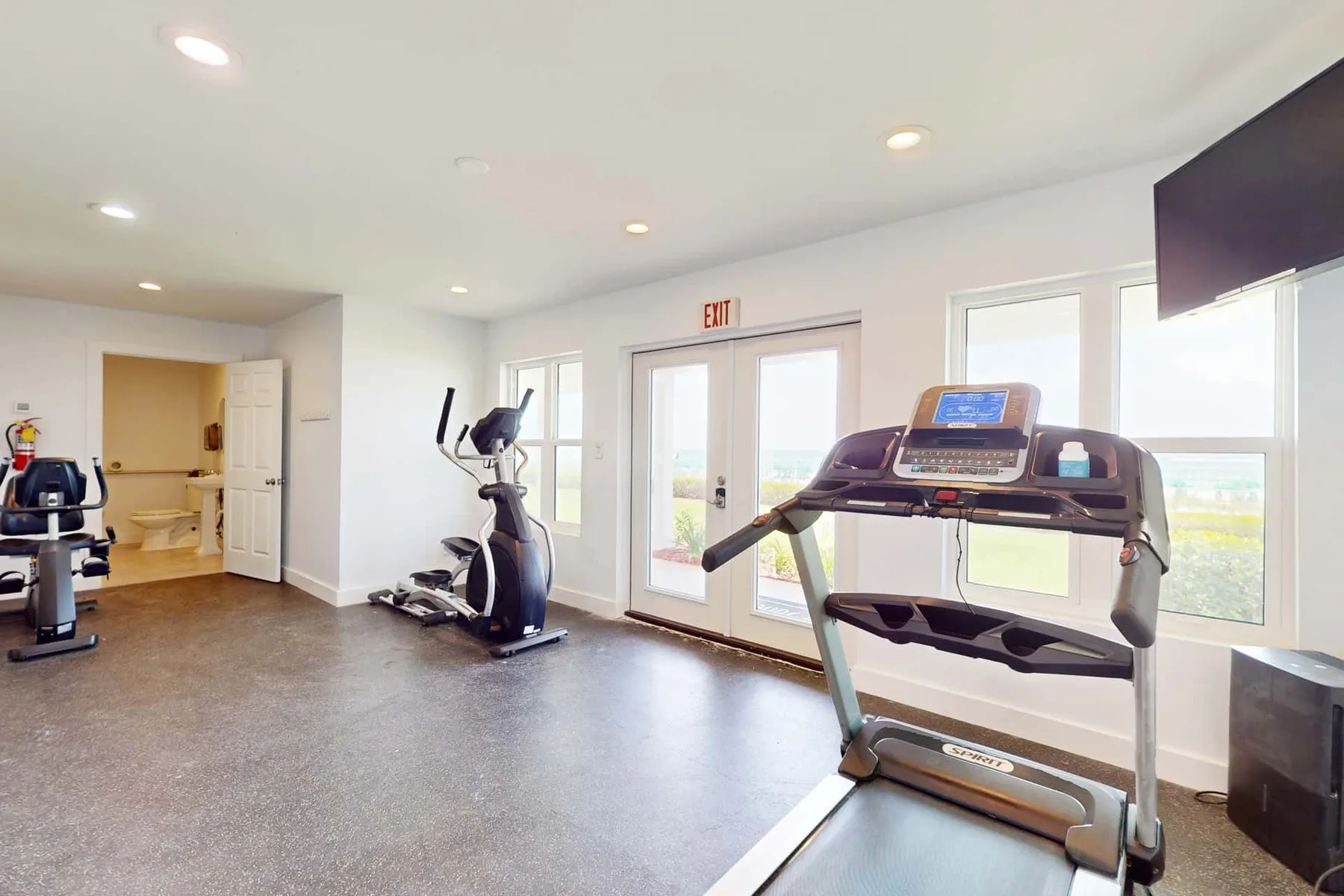 A bright fitness room with exercise machines and large windows.
