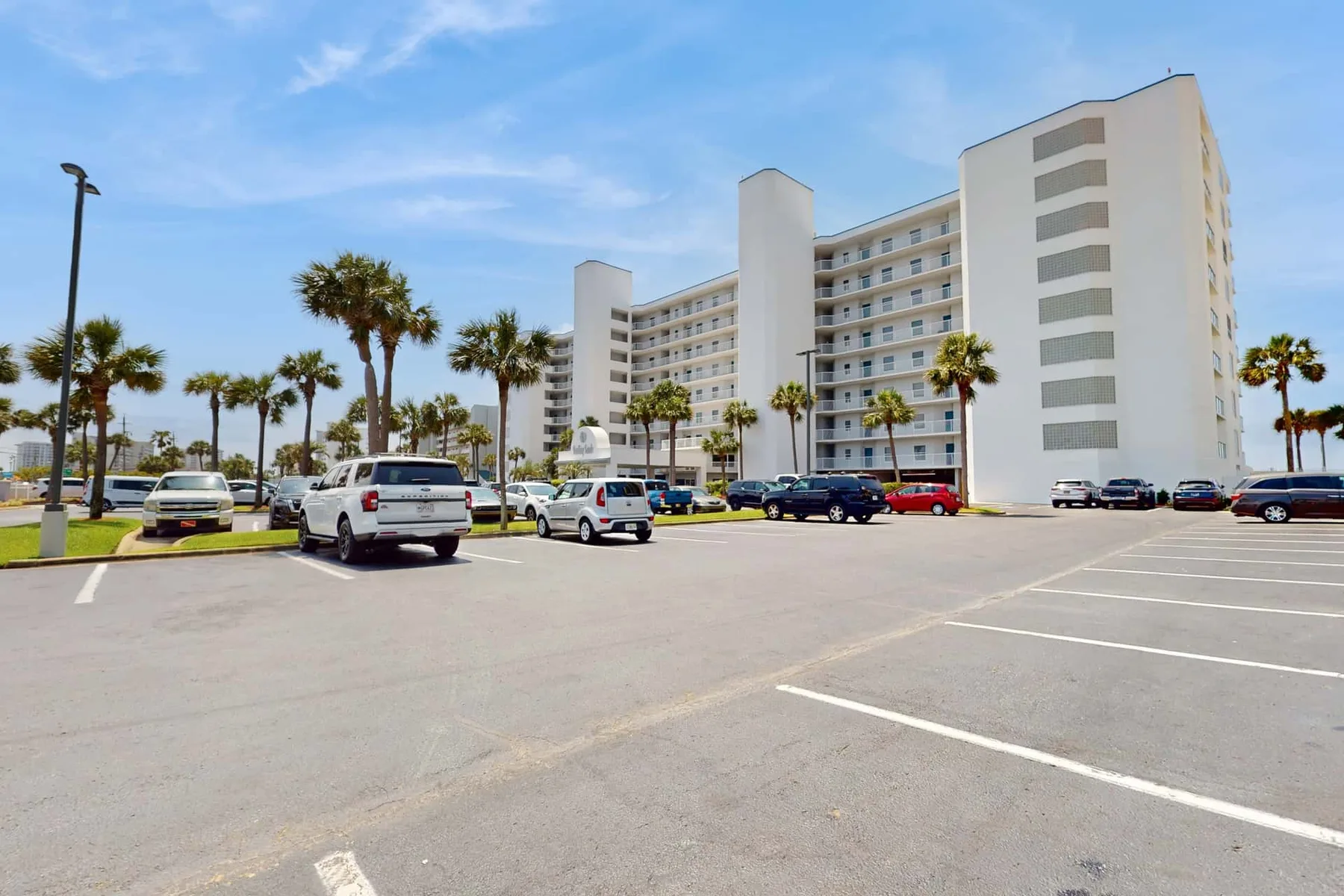 A white multi-story building overlooks a parking lot lined with palm trees and cars.